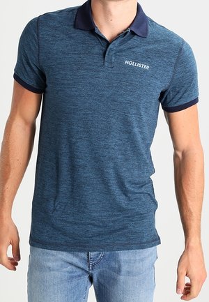Dunkelblaues Poloshirt mit kurzen Ärmeln, geripptem marineblauem Kragen und Bündchen, mit weißem Logo auf der linken Brust. Glatte Stoffstruktur.