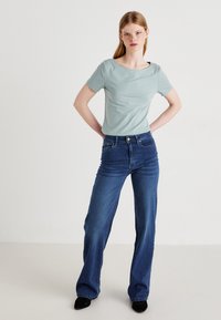 Top scurt de culoare bleu deschis cu un decolteu larg, asociat cu blugi închiși la culoare, cu talie înaltă. Modelul stă cu mâinile pe șolduri.
