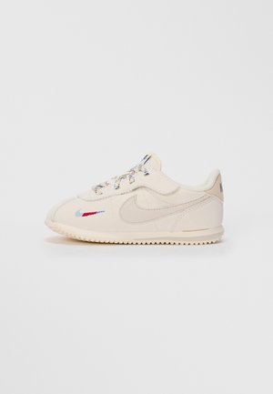 Baskets Nike blanches cassées avec semelle texturée, logo swoosh rouge et bleu brodé sur le côté, et lacets à motifs sur un fond uni.