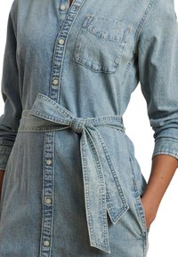 Light blue denim shirt dress with long sleeves, a collar, front buttons, a chest pocket, and a tied waist belt. Textured fabric finish.