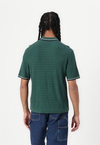 Polo shirt verde lavorato a maglia con un motivo testurizzato, maniche corte e colletto e polsini in un verde scuro a contrasto. Indossato con pantaloni cargo blu.