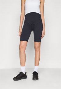 Shorts da moto in gravidanza neri con una vita alta senza cuciture, realizzati in tessuto elastico, abbinati a una canotta bianca e a scarpe da ginnastica nere.