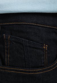 Jeans de mezclilla oscura con un bolsillo trasero visible, con costuras naranjas en contraste y una textura suave, llevados debajo de una camisa azul clara.