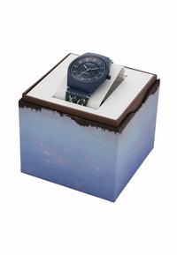 Blå klocka med oktagonal boett, texturerad mörk urtavla och stora siffror, tillsammans med ett vävt flerfärgat armband, förpackad i en naturskön låda.