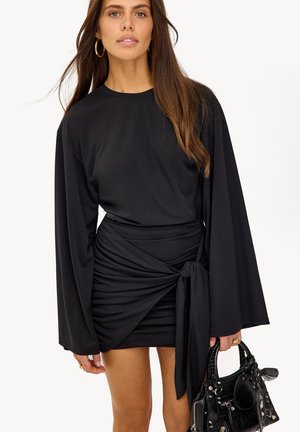 Femme portant une robe noire à manches longues avec une jupe asymétrique nouée, tenant un sac à main noir, aux longs cheveux bruns et boucles d'oreilles créoles en or.