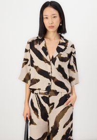 Chemise à manches courtes, à col, de couleur beige avec des rayures abstraites noires et brunes. Possède une poche sur le devant et une fermeture à boutons. Associée à un pantalon à motifs.