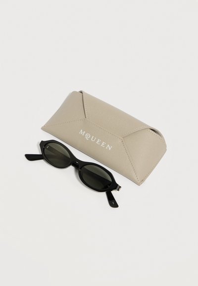 Μαύρα οβάλ γυαλιά ηλίου με σκοτεινούς φακούς δίπλα σε μπεζ δερμάτινη θήκη με ανάγλυφο το όνομα "McQueen" σε λευκά γράμματα.
