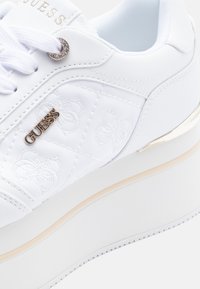 Sneaker platform bianco con lacci bianchi, logo dorato "GUESS", motivi cuciti e un piccolo occhietto di strass sul pannello laterale.