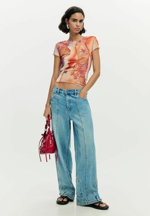 Jeune femme aux cheveux courts et foncés porte un haut coloré ajusté à manches courtes, un jean large bleu, des sandales noires, et tient un sac à main rouge.