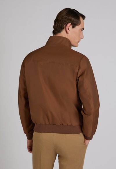 Uomo di spalle che indossa una giacca marrone con vita elastica e pantaloni beige, in piedi davanti a uno sfondo grigio chiaro uniforme.
