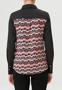 Chemise à manches longues avec un devant noir et un dos à motifs présentant des formes géométriques en rose, rouge et noir. Tissu texturé avec des accents de boutons.