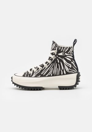 Sneaker alta con motivo astratto bianco e nero, suola piattaforma bianca spessa, suola esterna nera dentata e lacci bianchi.