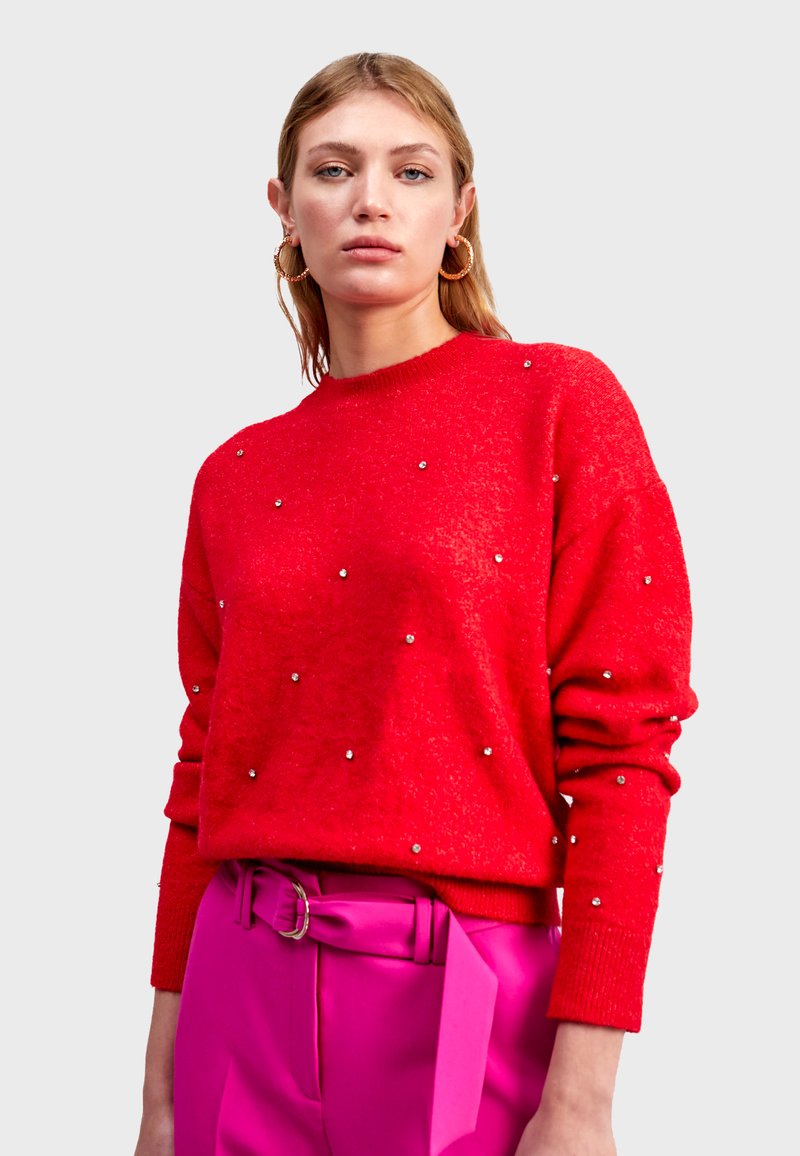 Un pull rouge avec une texture douce, orné de clous en argent, comportant un col rond et des manches longues, associé à un pantalon rose taille haute.