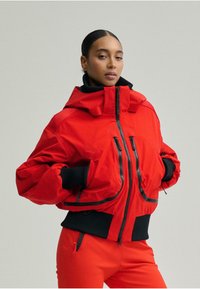 Piros steppelt dzseki magas gallérral, cipzáras záródással és elasztikus mandzsettákkal. Rövidített kialakítású, két cipzáras elülső zsebbel.