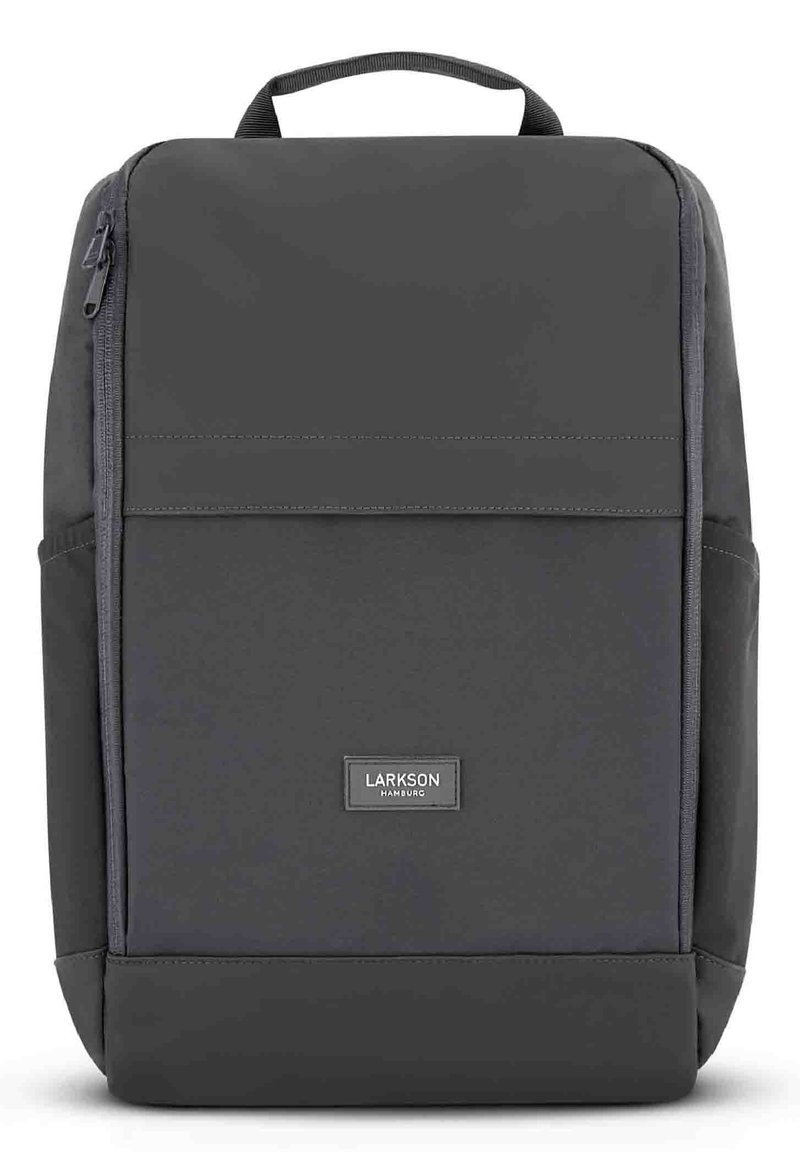 Zaino grigio con tessuto liscio, forma rettangolare, maniglia superiore, tasca con zip frontale e un discreto patch con logo nella parte inferiore.