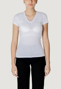 T-shirt bianco a V in cotone leggero, con maniche corte e un logo discreto sul petto. Abbinato a pantaloni neri aderenti.