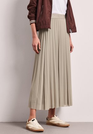 Personne portant une jupe maxi plissée beige, des baskets blanches, un haut blanc et une veste zippée bordeaux, debout contre un mur clair.