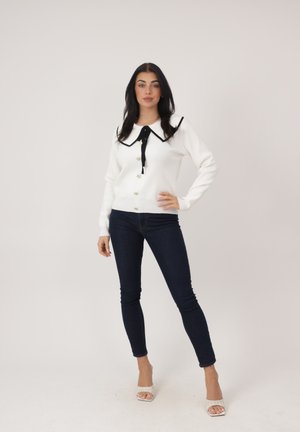 Kvinde stående med hånden på hoften iført hvid sweater med sort sløjfe ved kraven, mørke skinny jeans og hvide åbne hæle på ensfarvet baggrund.