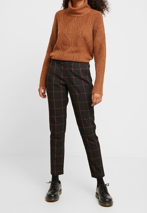 Vrouw draagt een bruine kabelgebreide coltrui, zwarte broek met rood ruitpatroon, zwarte sokken en zwarte veterschoenen, staand tegen een witte achtergrond.