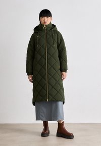 Gequiltede groene pufferjas met een hoge kraag, gouden rits en verstelbare koordjes. Gecombineerd met een denimrok en bruine enkelboots.