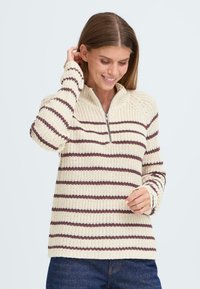 Strickpullover mit einem Viertelreißverschluss, ausgestattet mit cremefarbenen und dunkelbraunen horizontalen Streifen. Strukturiertes Gewebe mit gerippten Bündchen und Saum.