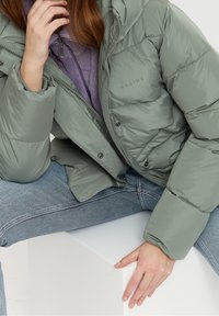 Lyst grøn pufferjakke med hætte, der har en blank tekstur, trykknapper og mærkenavnet "MAZINE" trykt på forsiden.