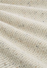 Close-up van crème-kleurige, gestructureerde breistof met een wafelweefpatroon en kleine bruine en zwarte vlekjes verspreid over de stof.