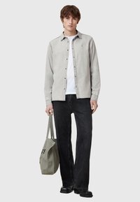 Helles graues Hemd mit Knöpfen, darunter ein weißes T-Shirt, schwarze Jeans und schwarze Schuhe. Hält eine graue Tragetasche mit bedrucktem Text.