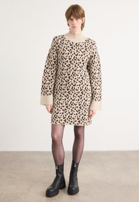 Vestido de malha em bege com estampa de leopardo em preto e castanho, apresentando um decote alto e mangas largas. Usado com botas pretas de fecho.