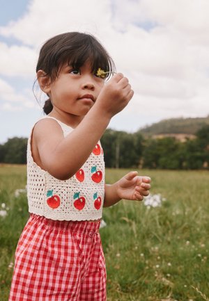 Jeune fille portant un haut à motifs et un pantalon rouge à carreaux tient une fleur jaune dans un champ vert avec des collines et des arbres en arrière-plan.