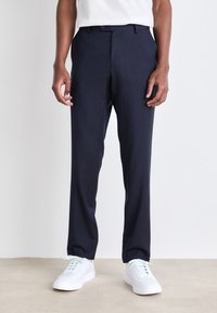 Pantaloni navy su misura in un tessuto leggero e liscio, con una vestibilità dritta e una piega sottile, abbinati a sneakers bianche.