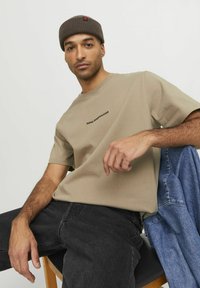 Bēšas kokvilnas T-krekls ar melnu tekstu "ROYAL DENIM DIVISION" uz krūtīm, kombinēts ar melnām džinsiem. Modelis valkā tumši brūnu adītu beanie cepuri.
