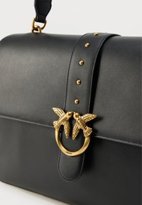 Μαύρη δερμάτινη τσάντα χειρός με λουράκι που έχει χρυσά καρφιά και χρυσό κούμπωμα σε σχήμα δύο πουλιών που φιλιούνται.