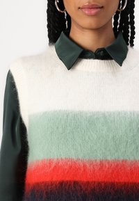 Sweter dzianinowy w kolorach ecru, zielonym, czerwonym i granatowym w paski; miękka faktura; nałożony na ciemnozieloną satynową koszulę z wyraźnym kołnierzykiem.