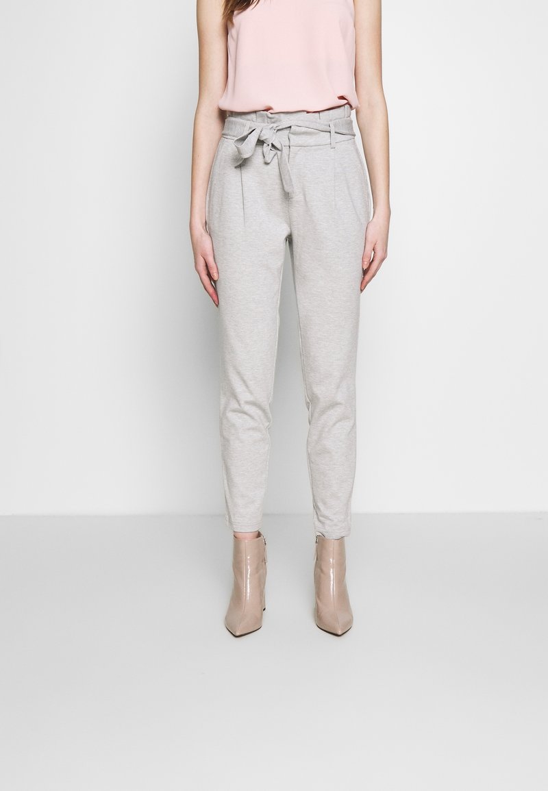 Femme portant un pantalon paperbag taille haute gris clair avec une ceinture en tissu nouée en nœud, un top sans manches rose blush et des bottines beiges.
