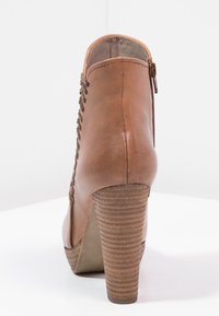 Bottine en cuir marron avec un talon haut, présentant des côtés texturés et une fermeture éclair à l'arrière. Des lacets décoratifs montent le long du côté.