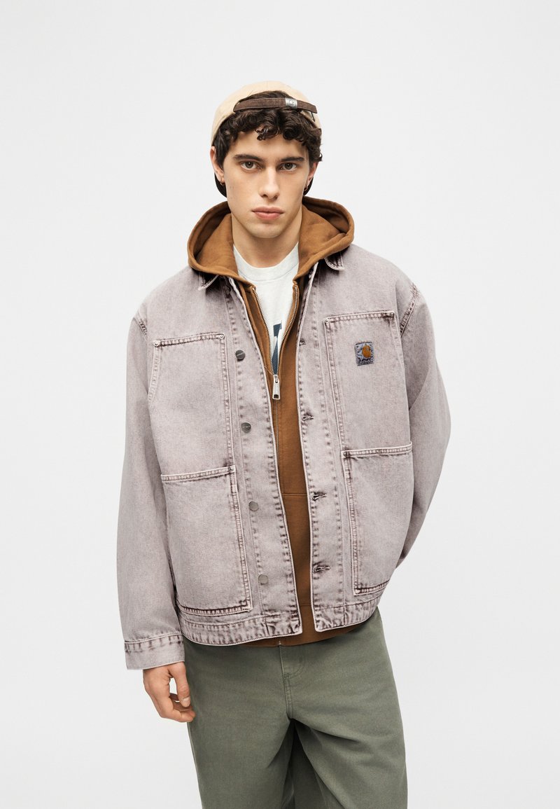 Jeune homme portant une casquette beige à l'envers, un sweat à capuche marron, une veste en jean délavée claire, et un pantalon vert, debout devant un fond uni.