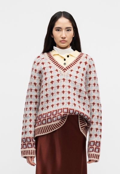 Femme portant un pull tricoté à motifs multicolores sur une chemise jaune à col et une jupe marron, debout devant un fond blanc.