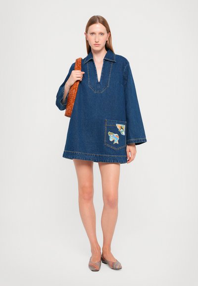 Denim dress with long sleeves, deep v-neck, and patch pocket featuring colorful embroidered details. Worn with a braided bag and flat shoes.