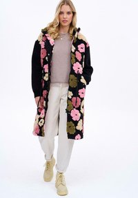 Dlhý čierny kvetinový kabát s ružovými, krémovými a zelenými kvetmi; mäkký materiál; pod ním krémový sveter; svetlé nohavice; béžové topánky.