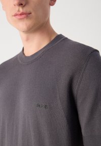 Szürke kötött pulóver bordás textúrával, kerek nyakkivágással és diszkrét fekete "BOSS" logóval a mellkason. Puha anyag, minimális design.