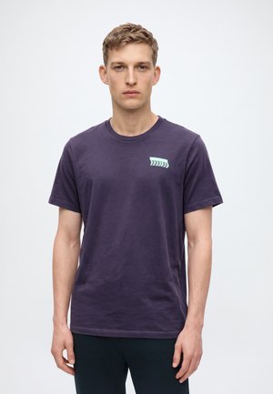 Un mannequin masculin se tient face à l’objectif devant un fond blanc uni. Il porte un T-shirt violet foncé avec un petit motif vert menthe et un pantalon foncé.