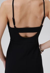 Schwarzer Jumpsuit mit dünnen Trägern, der einen Ausschnitt am Rücken, strukturierten Stoff und eine eng anliegende Silhouette aufweist. Sichtbare silberne Hardware-Akzente.