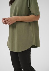 Camiseta verde oliva de corte relajado, cuello redondo, mangas cortas y dobladillo ligeramente curvado, combinada con pantalones negros.