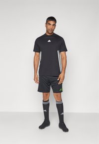 Svart trening T-skjorte med hvitt logo, svarte shorts med grønne detaljer, svarte sokker med hvite striper, og svarte fotballsko.