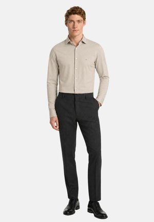 Uomo in piedi con camicia beige a maniche lunghe e pantaloni neri, con una mano in tasca, su sfondo bianco.