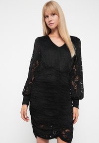 Μαύρο φόρεμα από δαντέλα με μακριά φουσκωτά μανίκια, διαθέτοντας V-ντεκολτέ, υφή και συγκεντρωμένες λεπτομέρειες στη μέση και το στρίφωμα.