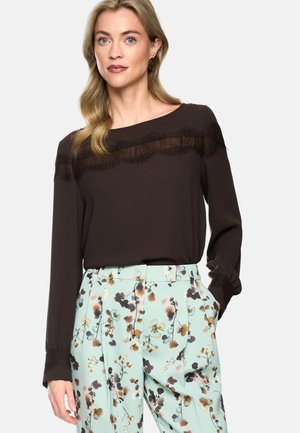 Frau trägt eine schwarze Langarmbluse mit Spitzendetails und hellblaue Hose mit floralem Muster, posiert mit einer Hand in der Tasche.