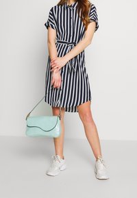 Abito button-up con strisce verticali blu navy e bianche, maniche corte, vita legata, abbinato a una borsa a tracolla verde menta con texture a coccodrillo e scarpe da ginnastica bianche.