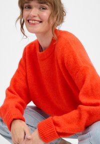 Živahno oranžna pletena pulover s krožnim izrezom in rebrastimi manšeti, izdelan iz mehkega in puhast materiala. Model je kombiniral s svetlo modrimi džinsi.
